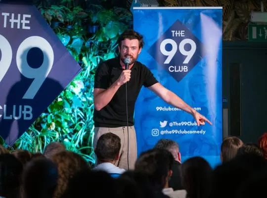 Man performing stand-up comedy on stage at The 99 Club with audience in foreground.