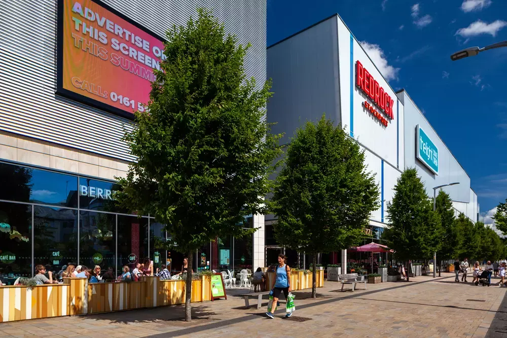 Outdoor shopping and leisure centre plaza with visitors, cafes, and modern retail buildings under blue sky.