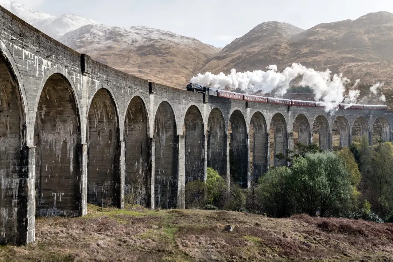 Steam train with red carriages crossing a stone railway viaduct beneath snow-capped mountains.