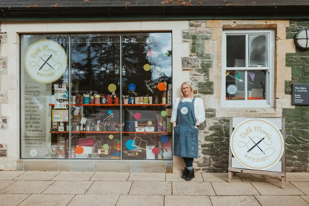 Woman wearing apron standing outside a creative workshop storefront with colourful displays in the windows.