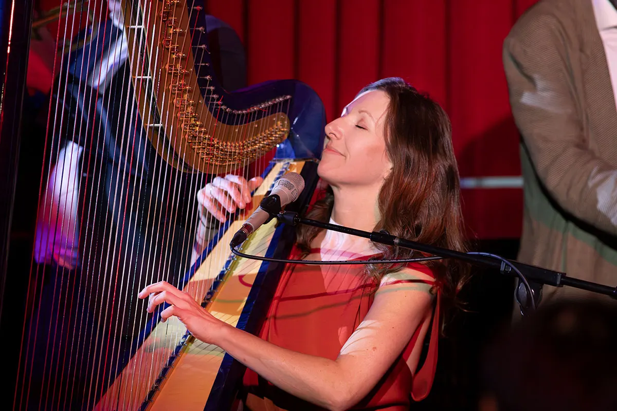 Musician performing with harp and singing into microphone on stage with red curtain backdrop.