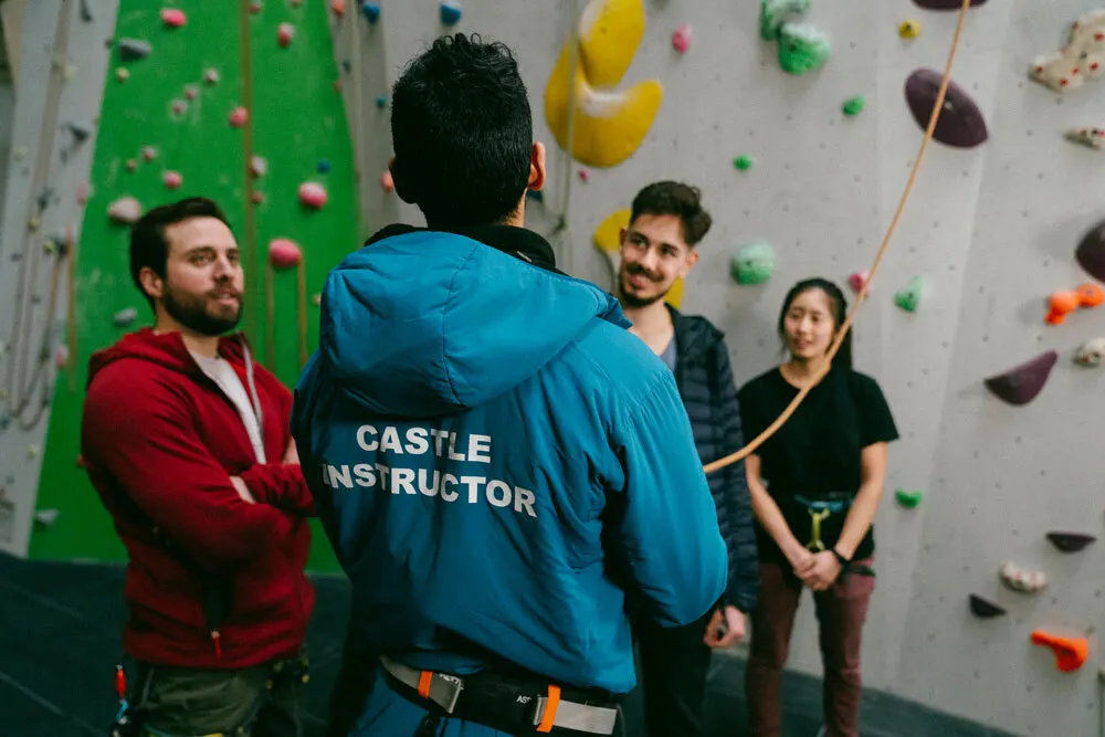 An instructor in a blue jacket talks to a group of people standing in front of a colourful indoor climbing wall.