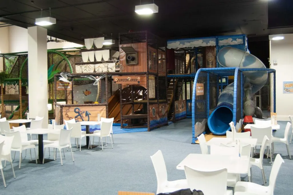 Indoor play area with large wooden and blue climbing structures, slides, and seating tables in foreground.