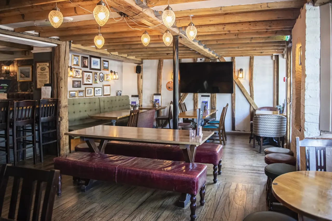 Spacious interior dining area with wooden beams, hanging pendant lights, burgundy leather benches, wooden tables and chairs, and framed photographs on walls.