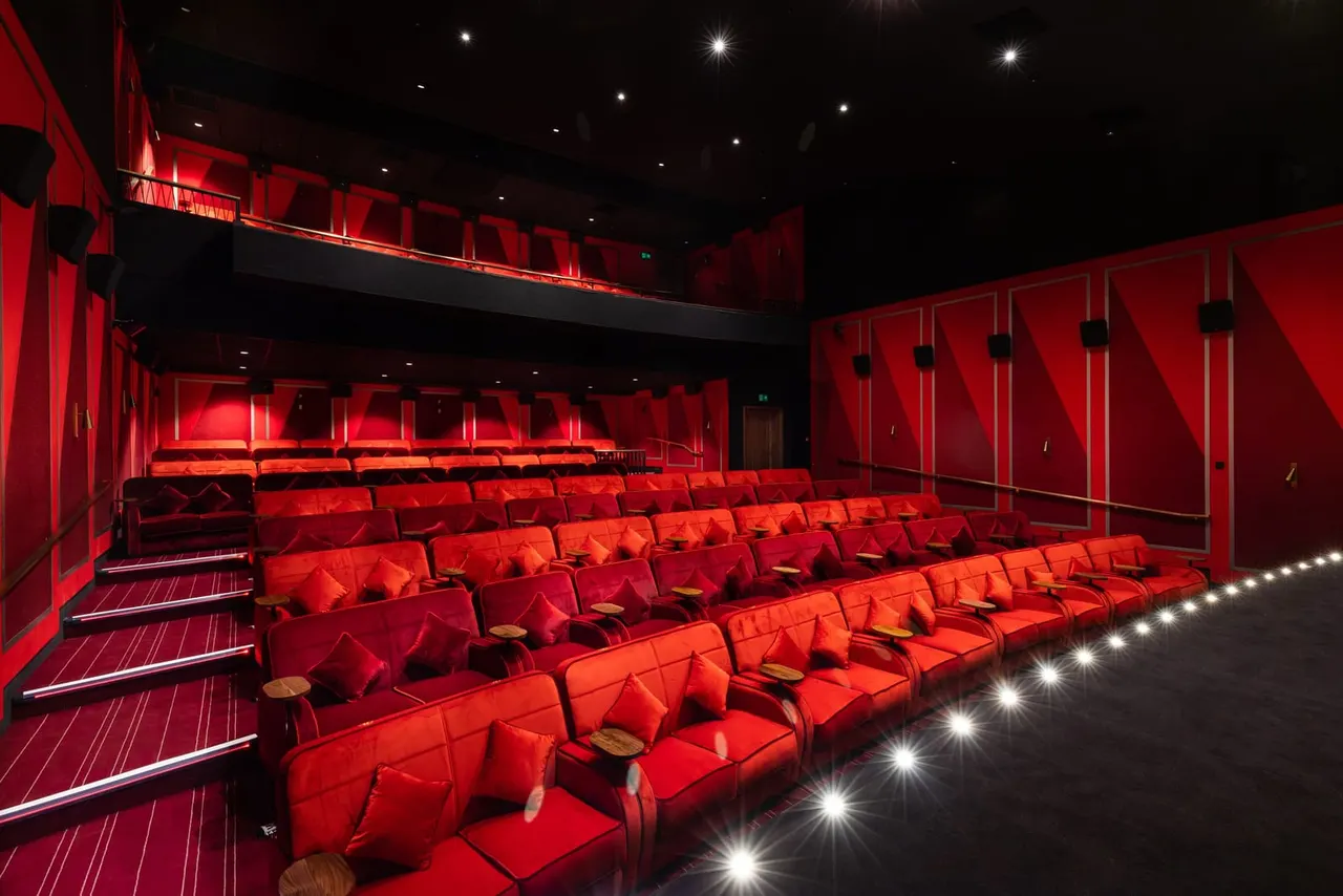 Empty cinema screen room with rows of red reclining seats, red walls, and illuminated floor lighting.