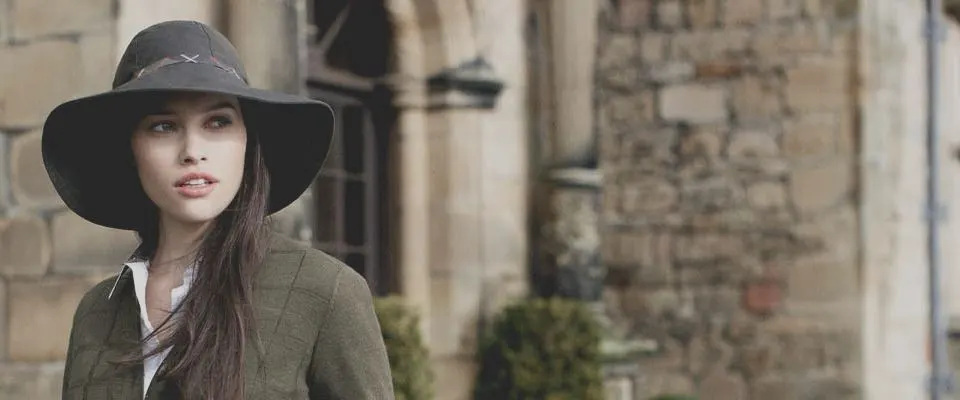 Woman wearing a wide-brimmed hat standing in front of stone castle walls.