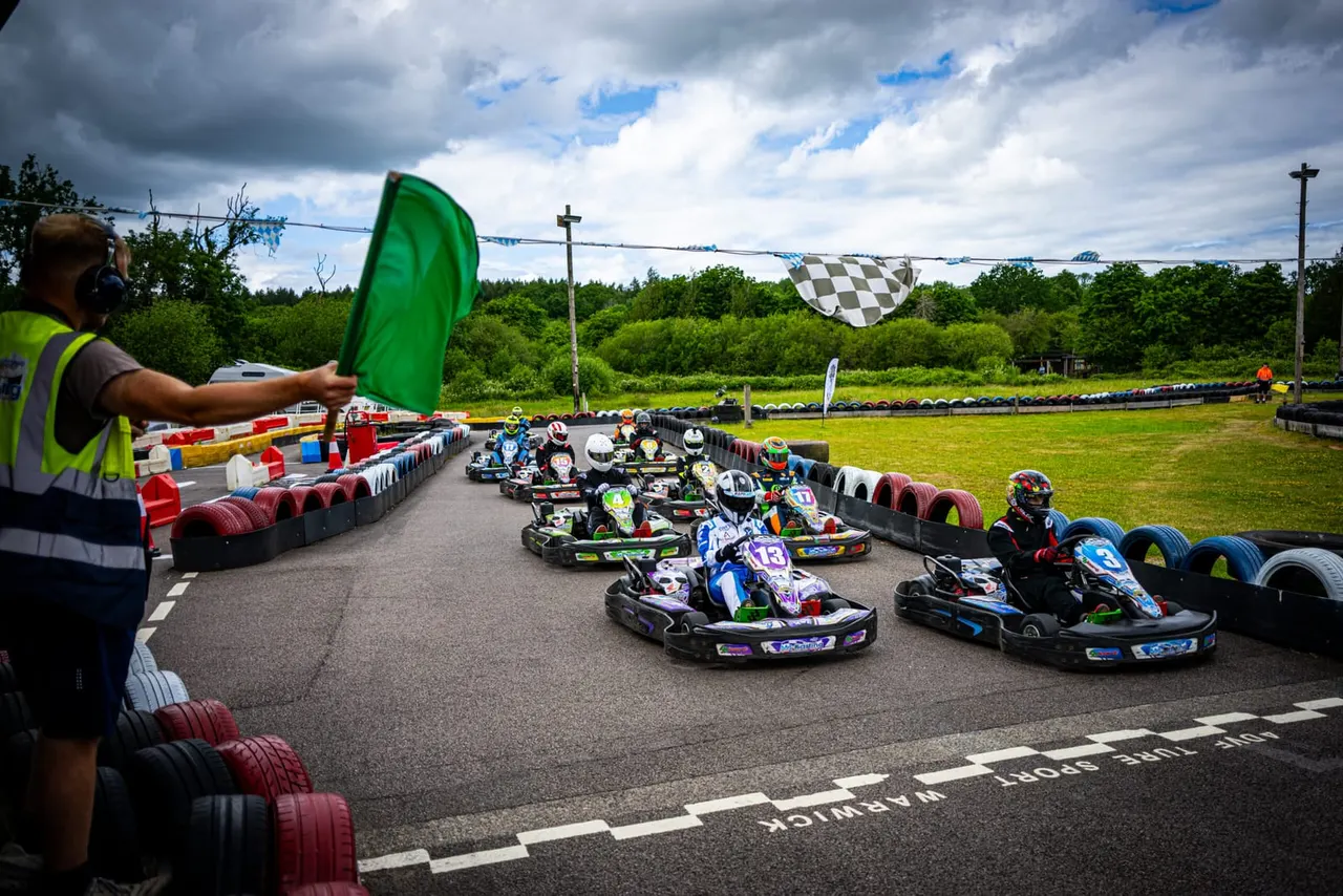 A marshal waving a green flag as go-karts race around an outdoor track lined with tyres and barriers.