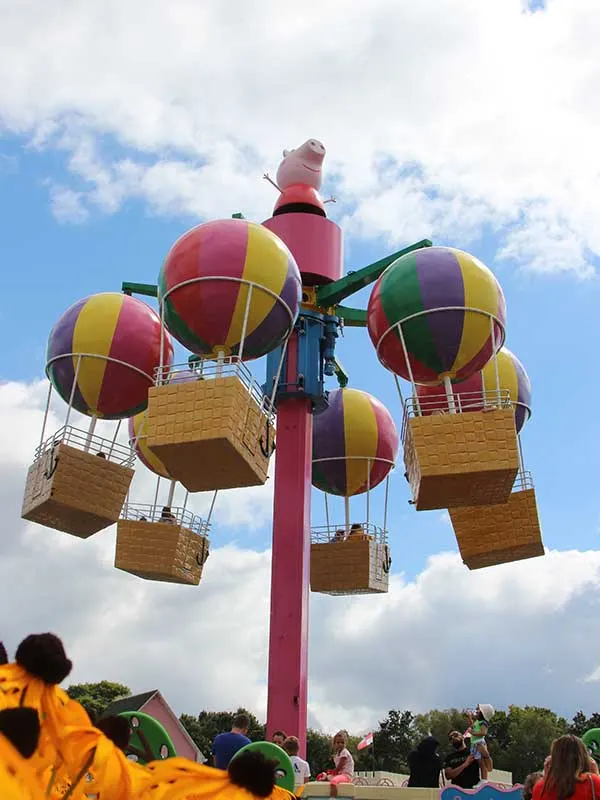 Colourful hot air balloon ride with wicker baskets suspended from a central pole against a cloudy sky, with visitors watching below.