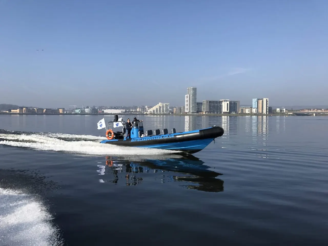 Blue and black speedboat with passengers approaching shore in shallow water, with cityscape and calm bay in background.