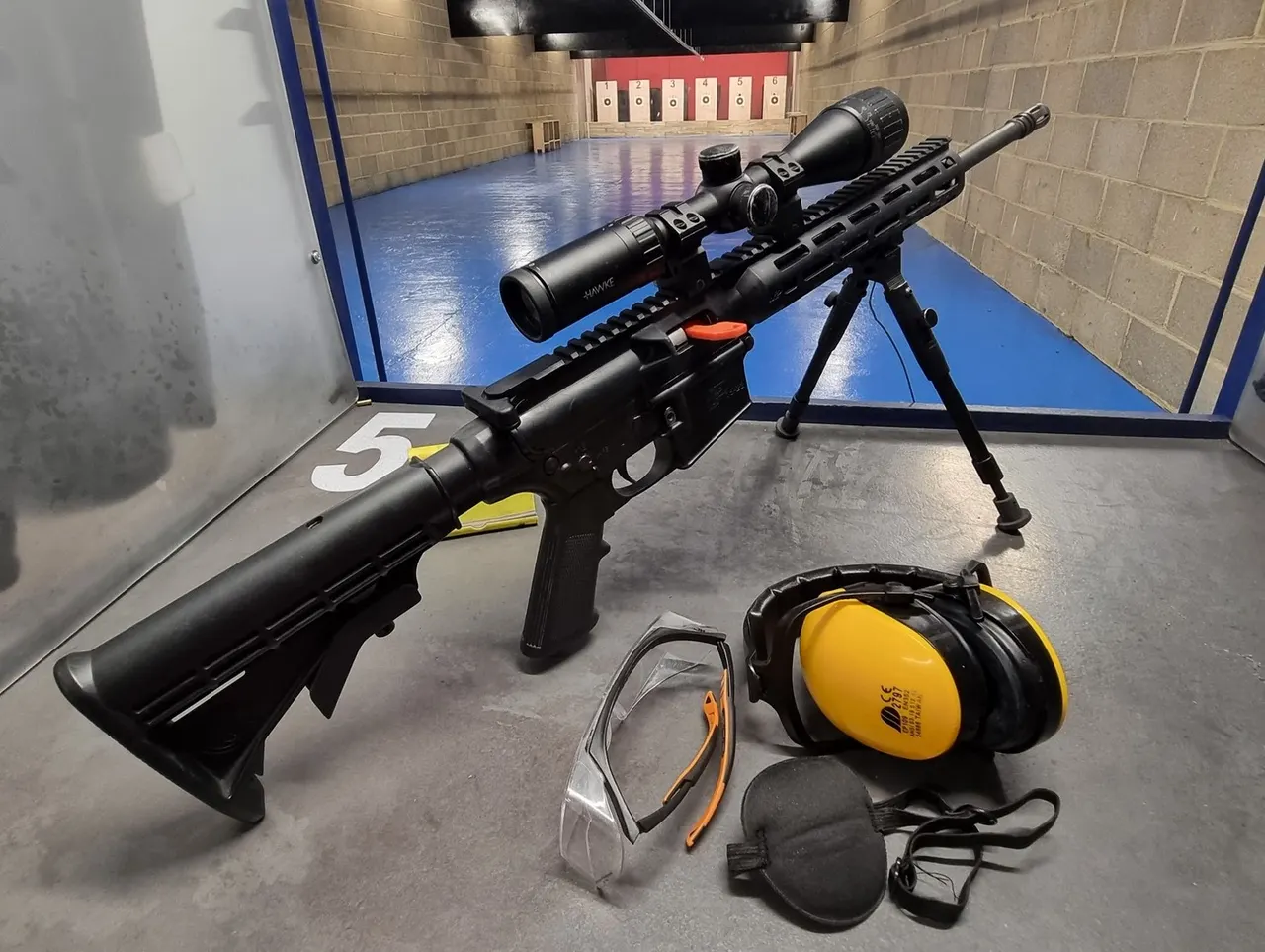 Rifle with scope and safety equipment displayed in an indoor shooting range with target boards visible in background.