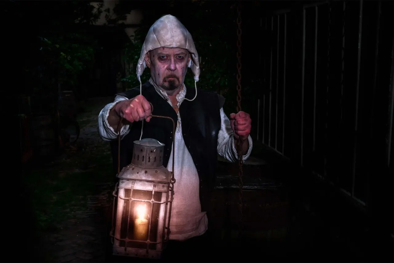 Man in Tudor costume holding a lit lantern, posing in character at night in a dark alleyway setting.