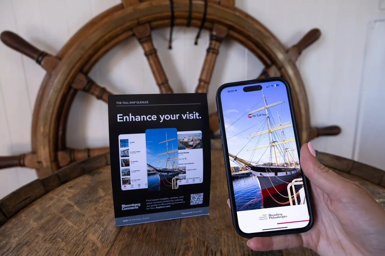 A hand holding a smartphone displaying the Tall Ship Glenlee app next to a promotional card, with a ship's wooden wheel in the background.