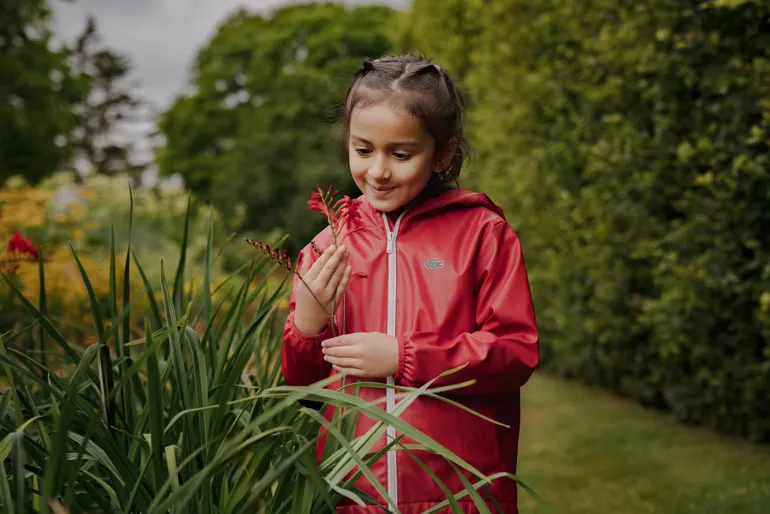 A young girl in a red jacket examines a red flower amongst plants in a garden setting.