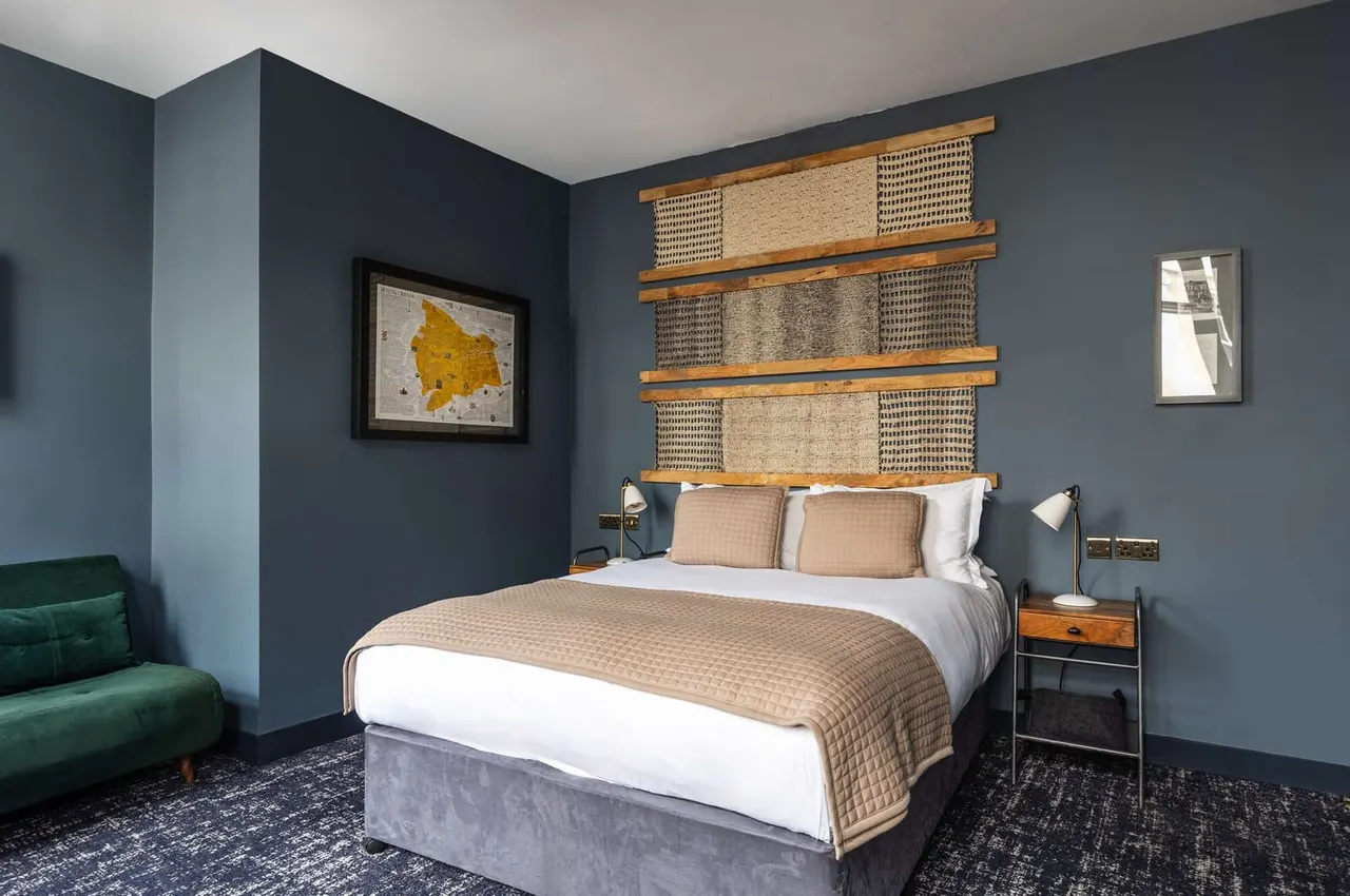 Modern hotel bedroom with double bed, woven wall panels above headboard, dark blue walls, framed map artwork and green velvet chair.