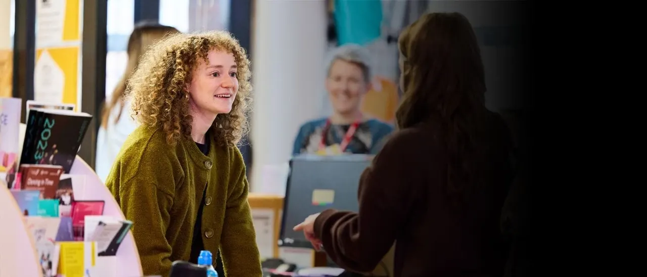 Young person with curly blonde hair smiling whilst speaking to another person at an indoor event or workshop.