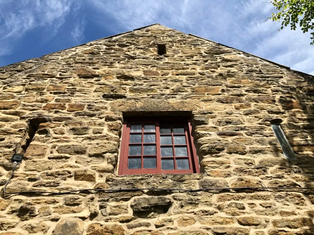 Stone gable end of historic mill building with red-framed window against blue sky.