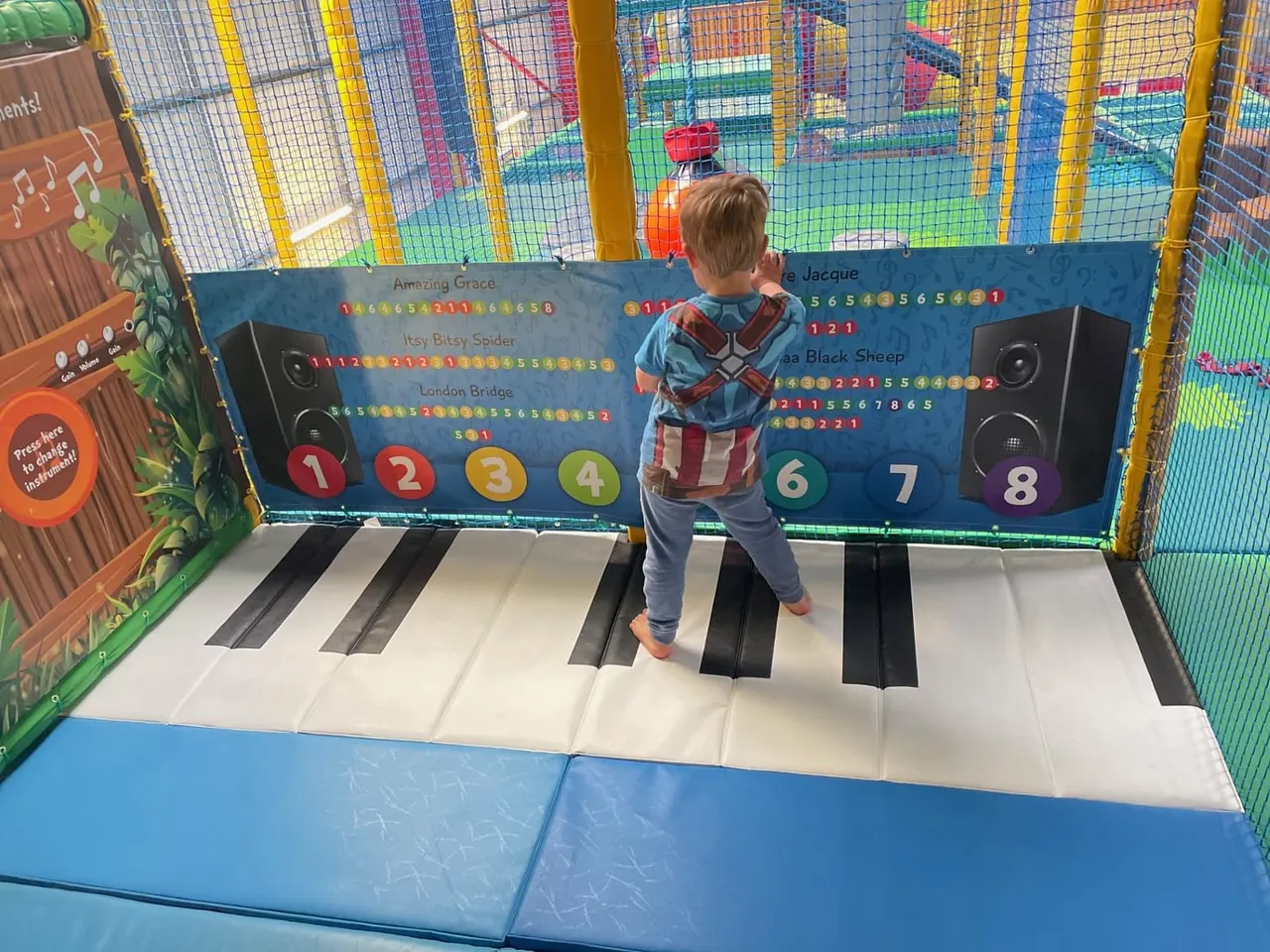 A young child plays on a giant interactive piano keyboard mat inside an indoor soft play area.