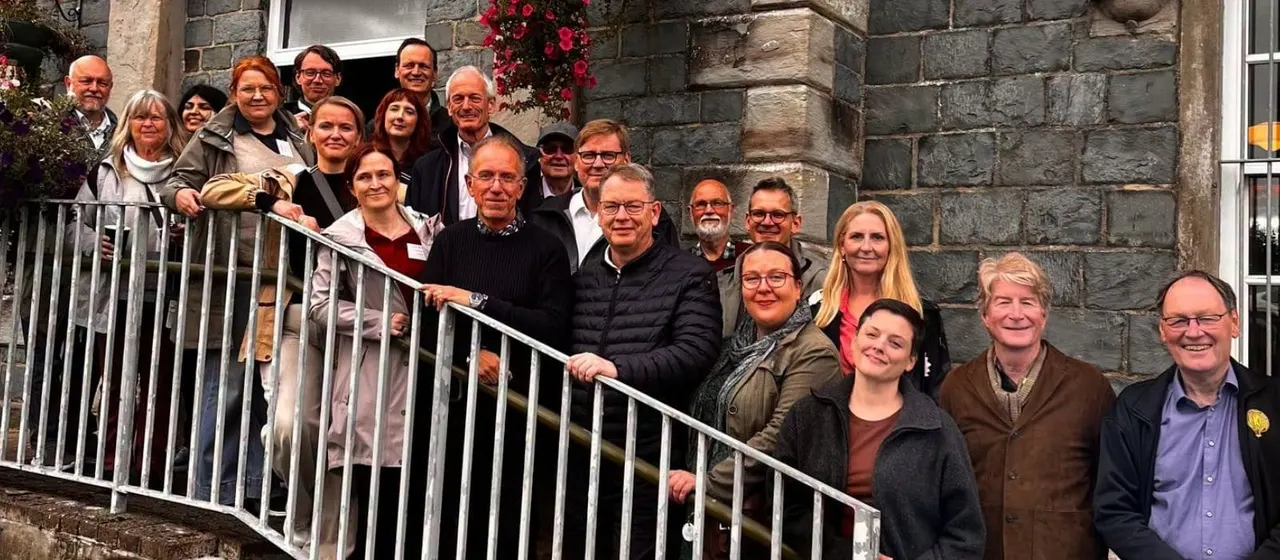 Group of approximately twenty-five adults posing together on a metal ramp outside a stone building with red flowers.