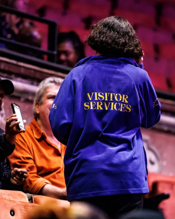 Visitor services staff member in blue jacket assists a guest in an indoor ice arena with red seating.