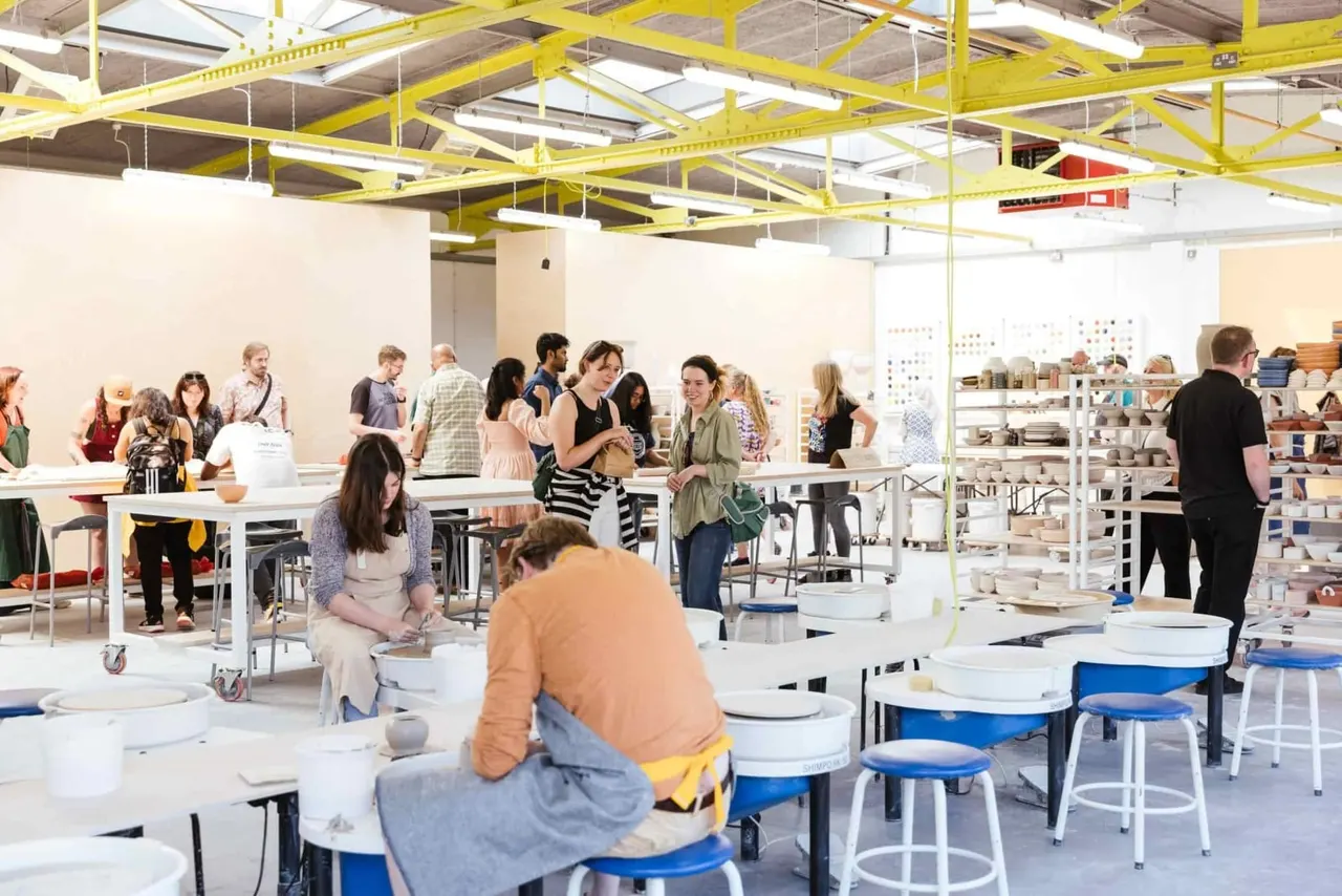 A pottery workshop with numerous visitors engaged in hands-on ceramic activities at various workstations and tables.