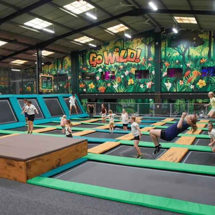 Children and adults bouncing on colorful trampolines in an indoor play facility with jungle-themed wall murals.