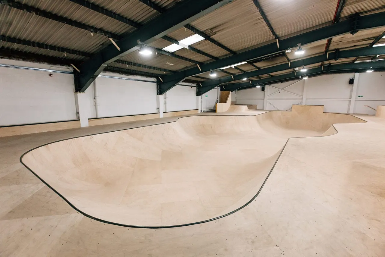 Empty indoor skate park with wooden ramps and bowls under industrial ceiling with LED lighting.
