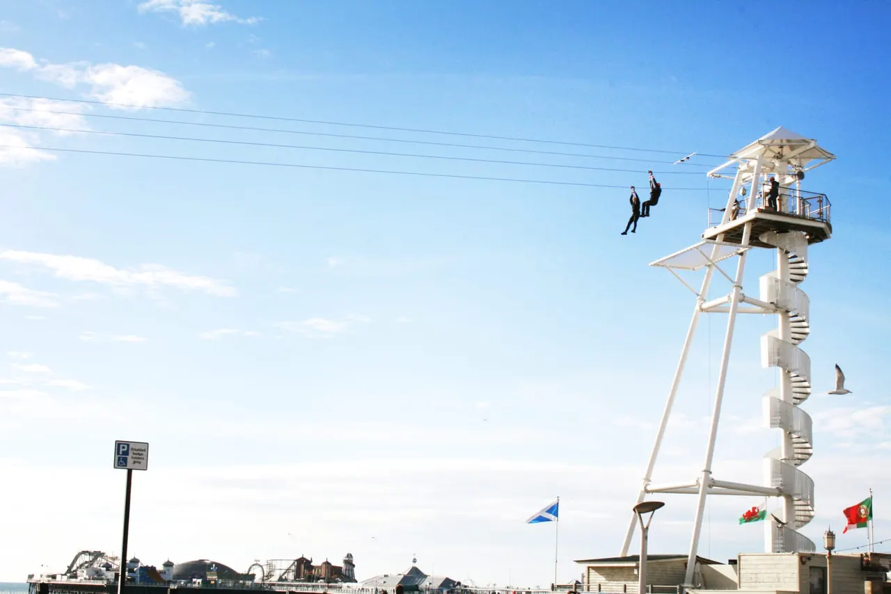 Two people zip-lining down cables from a tall white tower at a seaside leisure venue under blue sky.