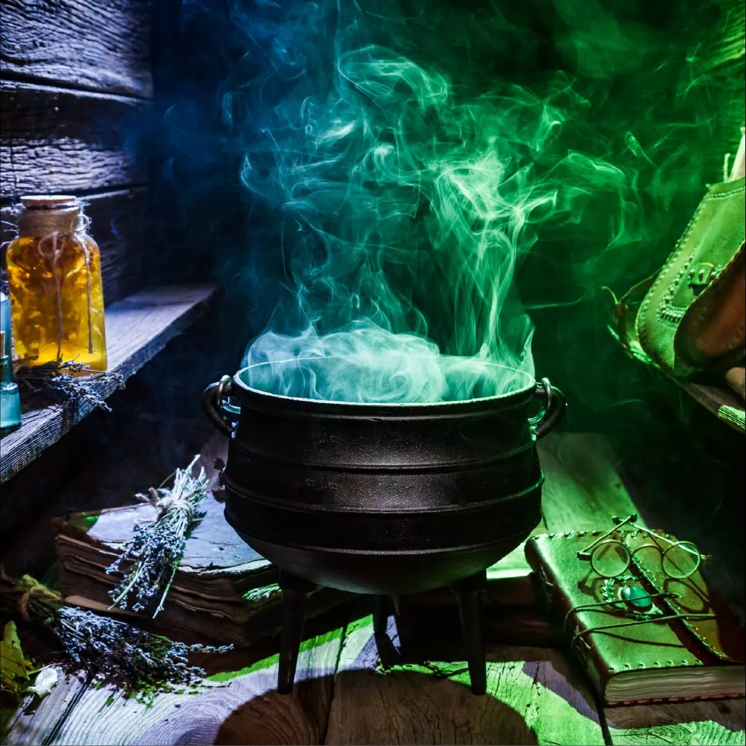 A black cauldron with green and blue glowing smoke billowing from it, surrounded by witchcraft props including bottles, books, and herbs on a wooden surface.