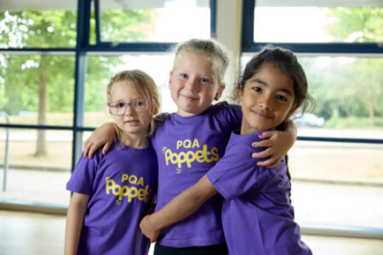 Three children wearing purple PQA Poppets t-shirts stand together smiling by a large window overlooking green grounds.