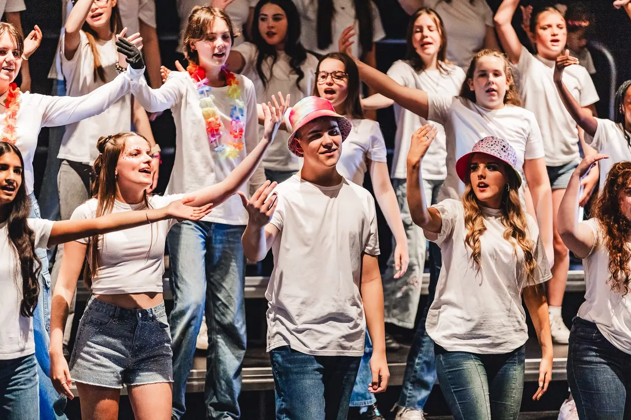 A group of young people in white t-shirts and jeans perform on stage together, singing and dancing with energetic gestures and expressions.