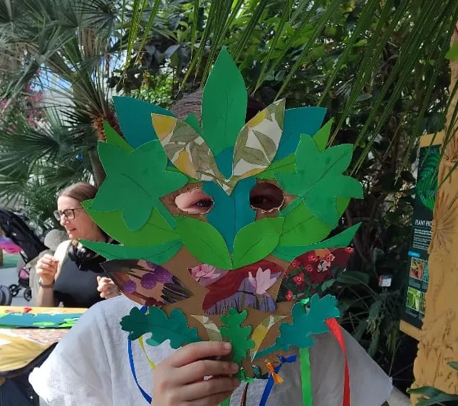 Child holding a large colourful paper leaf craft in front of their face in a greenhouse with tropical plants.
