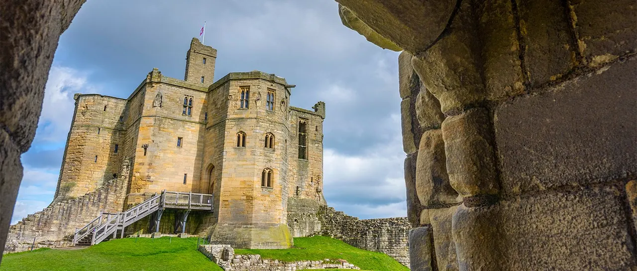 Stone castle with tower and flag viewed through ruined archway, set on grassy mound with wooden bridge entrance under blue sky.
