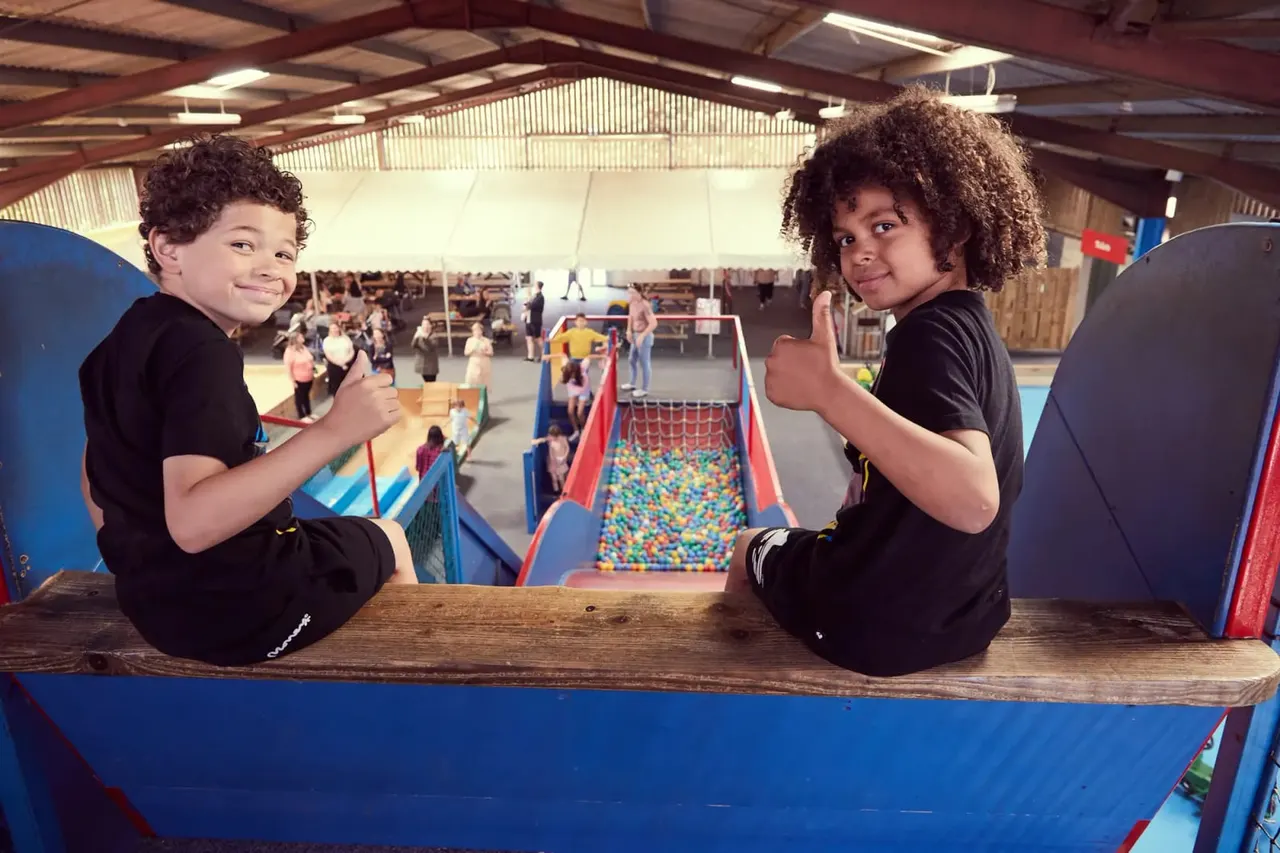 Two children giving thumbs up whilst sitting on a wooden beam overlooking a ball pit and soft play area.