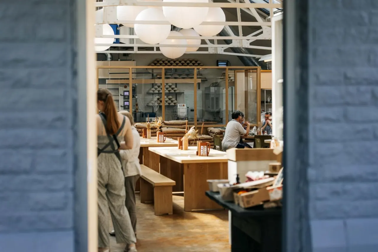 Modern café interior with wooden furniture, pendant lights, and customers seated at tables near glass-fronted kitchen area.