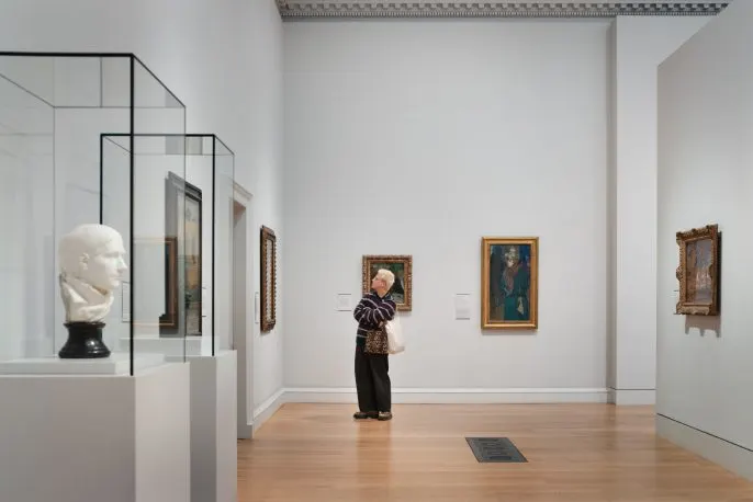 A person viewing framed paintings on white gallery walls, with a classical marble bust displayed in a glass case on the left.
