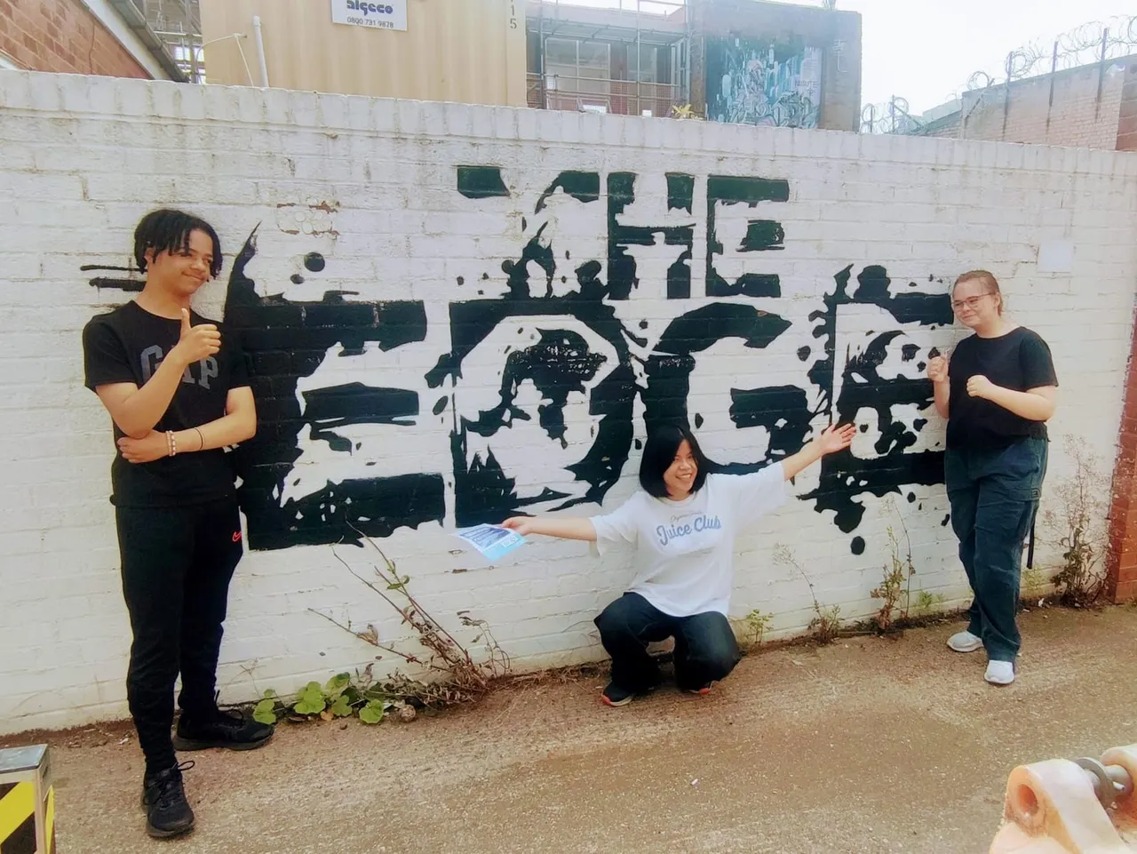 Three young people posing in front of a large black and white mural on a brick wall at an urban arts venue.