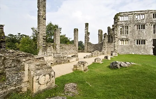 Ruins of a medieval castle with stone walls and pillars, a stone building with windows on the right, and green lawn in the foreground.
