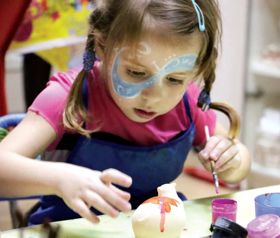 Child with face paint doing a craft activity at a table with paintbrushes and coloured containers.