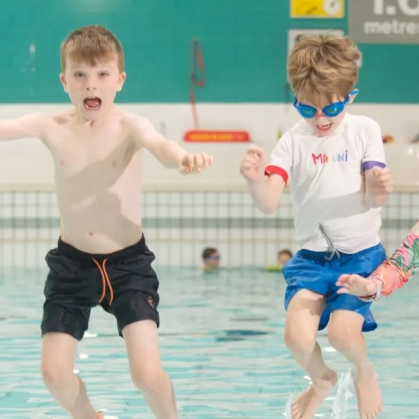 Two young children running into an indoor swimming pool, wearing goggles and swimming costumes.