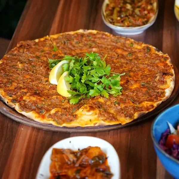 A round flatbread pizza topped with minced meat, herbs and lemon wedges on a wooden table.