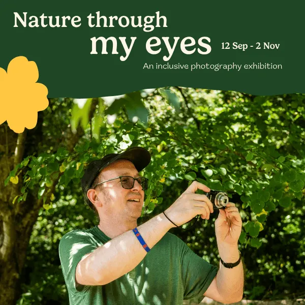 Man wearing glasses and green shirt holding a vintage camera whilst standing beneath lush green foliage.