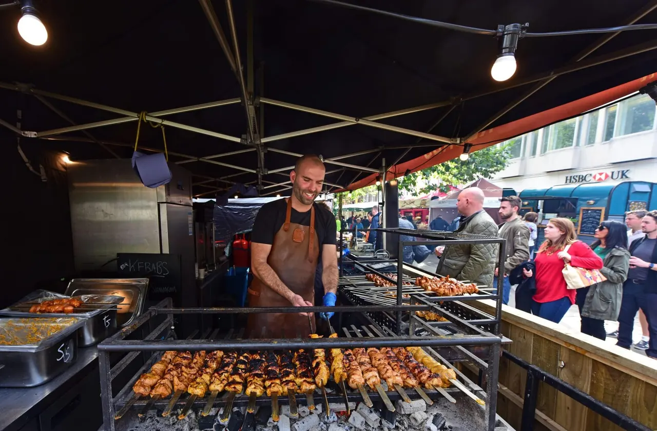 Man grilling meat skewers at an outdoor food stall whilst customers queue to order.