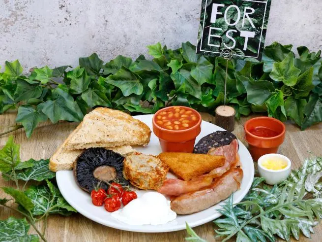 A full English breakfast with toast, sausages, bacon, baked beans, mushroom and tomato, surrounded by fresh greenery and a "Forest" sign.