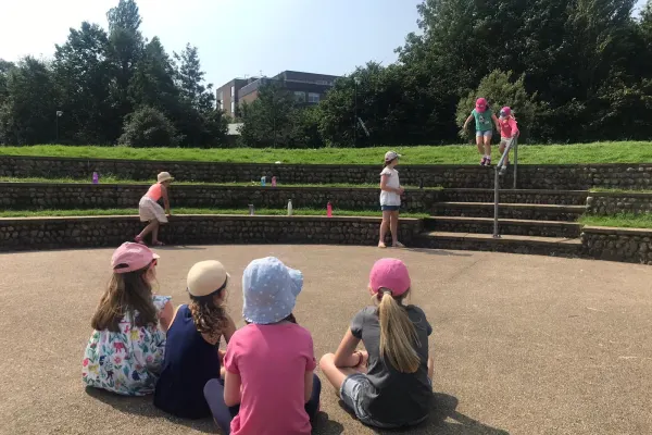 Children watching others play in a sandy arena surrounded by wooden fencing and green fields.