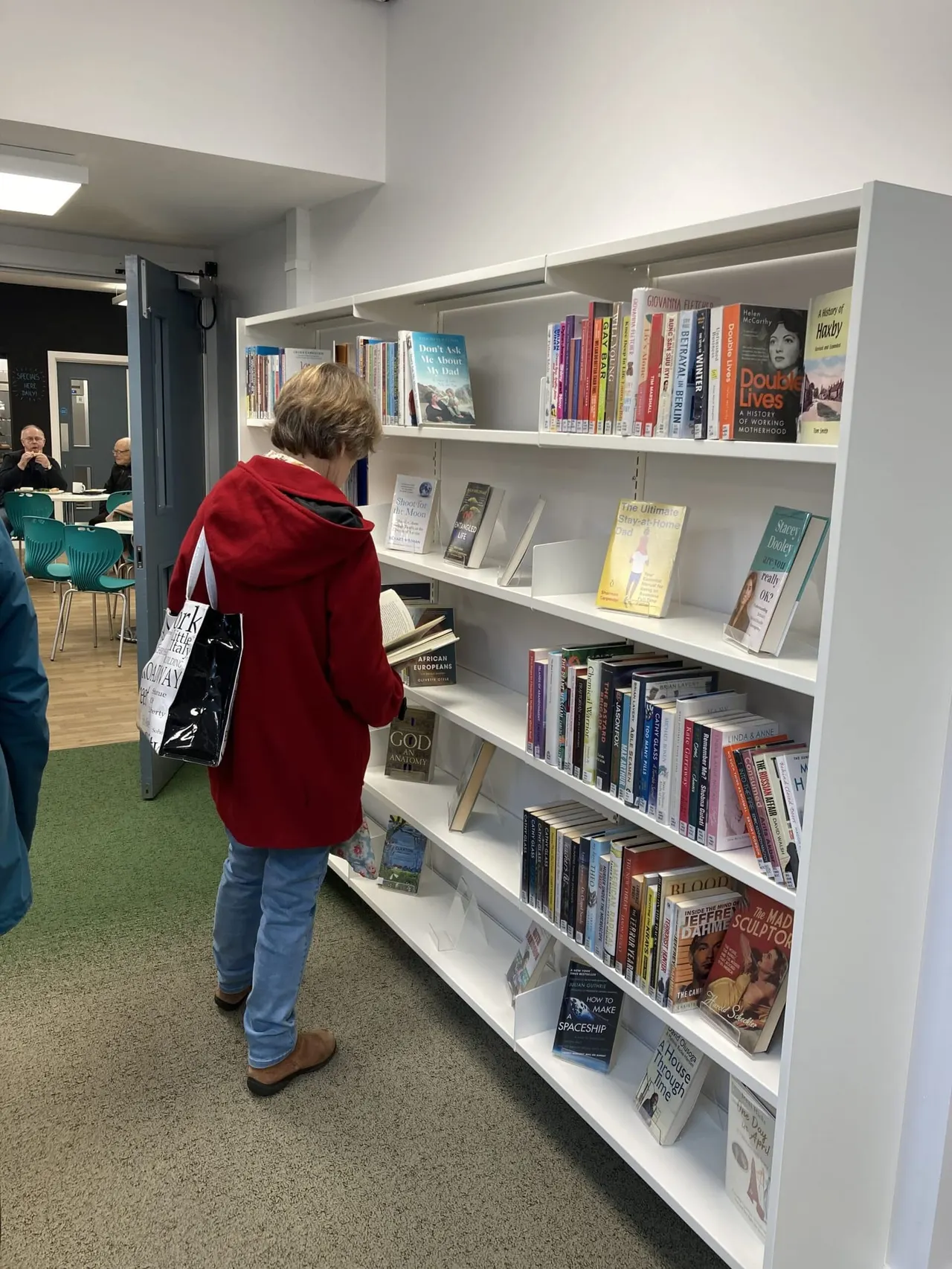 Person in red hoodie browsing books on white shelving unit in library, with café seating visible in background.