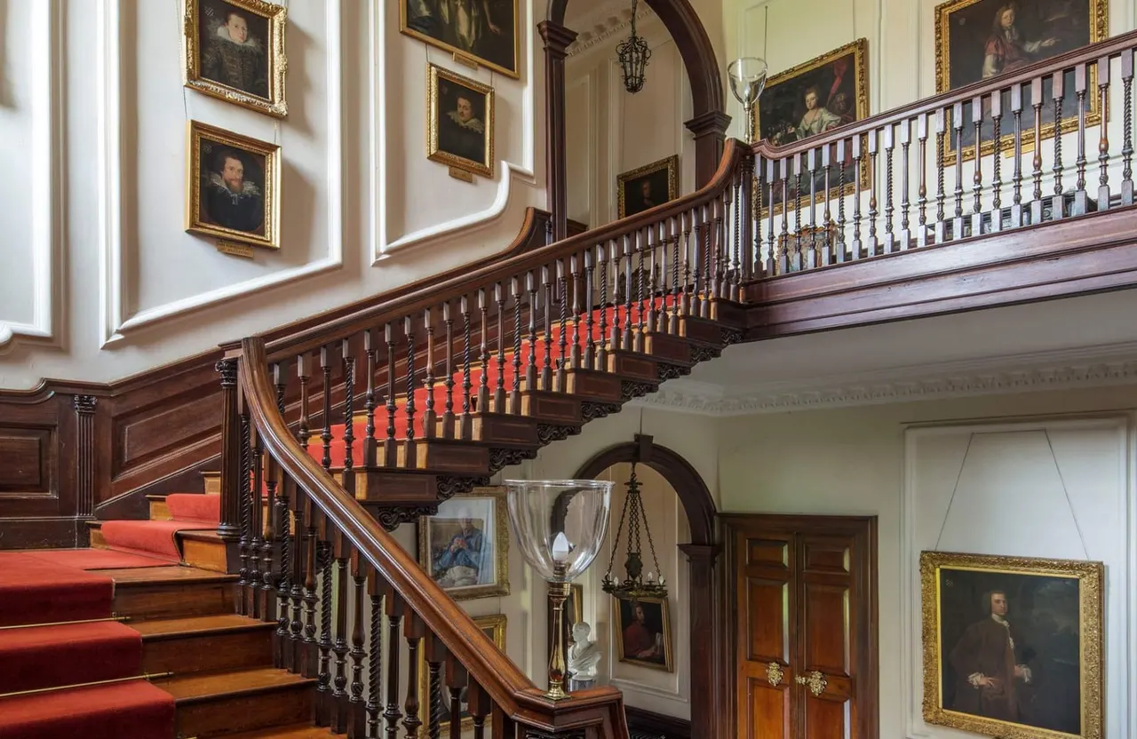 Grand wooden staircase with red carpet and ornate balustrade, lined with gilt-framed portraits and classical sculptures in an elegant period interior.