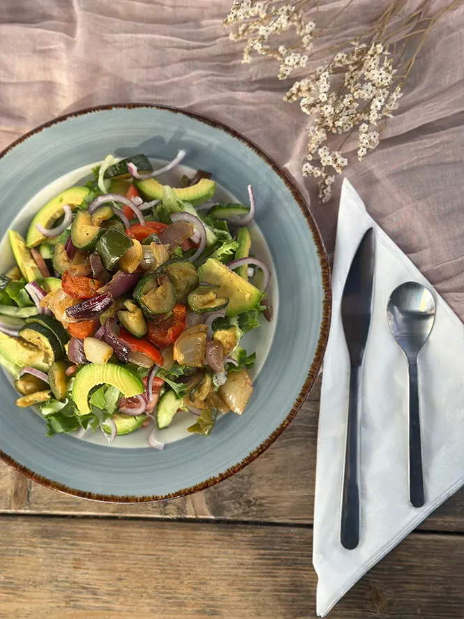 A blue-rimmed bowl of roasted vegetables and salad with cutlery and napkin on a wooden table.