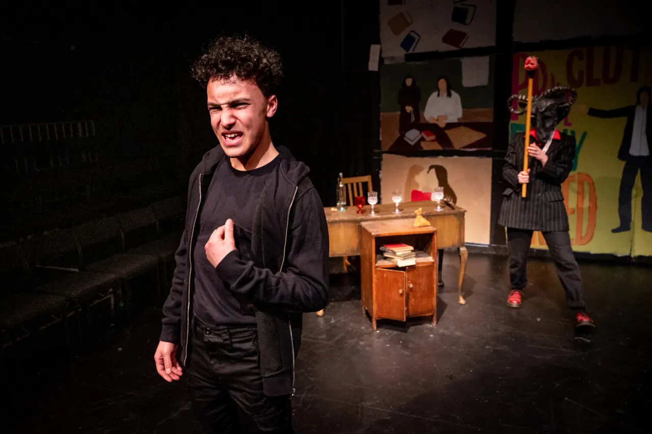 Young man performing on stage with other actors in a theatrical production set with furniture and props.