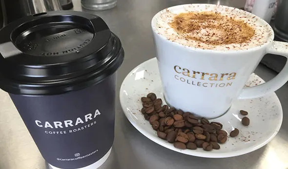A takeaway coffee cup and a cappuccino with cocoa powder beside coffee beans on a saucer.
