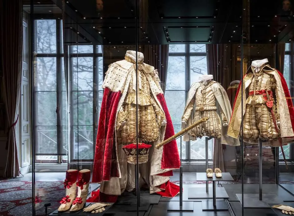 Historical royal ceremonial robes and regalia displayed in a museum gallery with large windows.